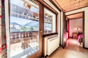 A room with a wooden floor, a radiator, a bed with a pink blanket, and a window with a view of buildings and mountains.