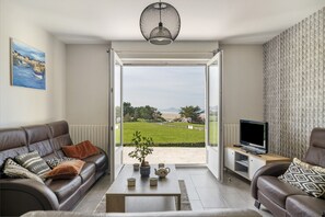 Uma sala de estar com sofá, mesa de centro e televisão, uma janela grande com vista para uma paisagem verde, e um quadro na parede.