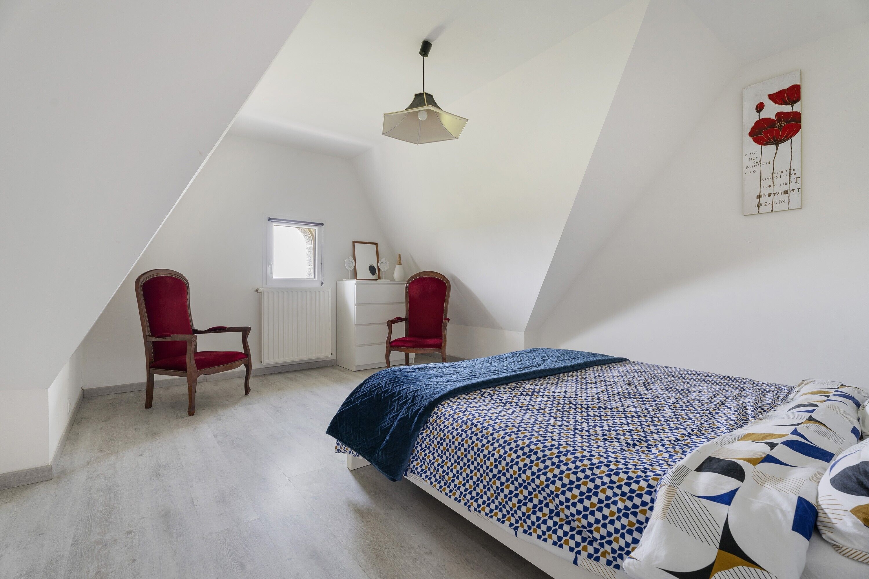 Une chambre avec un plafond en pente, un lit avec une literie à motifs, deux chaises rouges, un bureau avec une chaise et une œuvre d’art accrochée au mur.