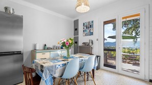 Een keuken met een koelkast, een eettafel met stoelen, een vaas met bloemen en uitzicht door een deur.