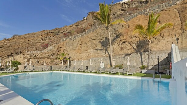 A swimming pool with a stone wall backdrop, lounge chairs, and palm trees. - Holiday Apartment 'Amazing' with Sea View, Shared Pool and Wi-Fi (Mogán)