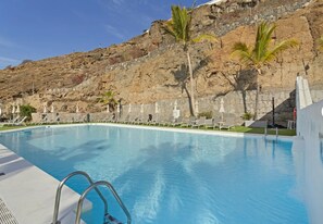 A swimming pool with a stone wall backdrop, lounge chairs, and palm trees. - Holiday Apartment 'Amazing' with Sea View, Shared Pool and Wi-Fi (Mogán)