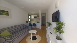 A living room with a sofa, a small round table, two chairs, a TV mounted on the wall, and a kitchen in the background. - Holiday Apartment 'Amazing' with Sea View, Shared Pool and Wi-Fi (Mogán)