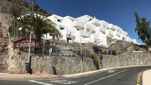 A white building with balconies, a stone wall, and a palm tree. - Holiday Apartment 'Amazing' with Sea View, Shared Pool and Wi-Fi (Mogán)