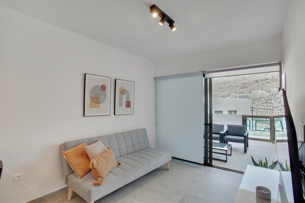 A modern living room with a grey sofa, two framed artworks, a sliding glass door leading to an outdoor seating area.