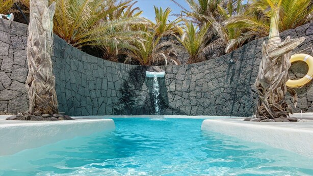 A pool with clear blue water, surrounded by a rocky wall and palm trees. - Vacation Villa 'El Cactus' with Shared Pool and Wi-Fi (Teguise)