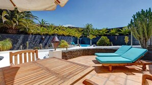 A wooden deck with a table and two lounge chairs, a swimming pool, and a stone wall with tropical plants. - Vacation Villa 'El Cactus' with Shared Pool and Wi-Fi (Teguise)