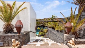 A pathway leading to a pool with stone walls and sculptures, surrounded by tropical plants. - Vacation Villa 'El Cactus' with Shared Pool and Wi-Fi (Teguise)