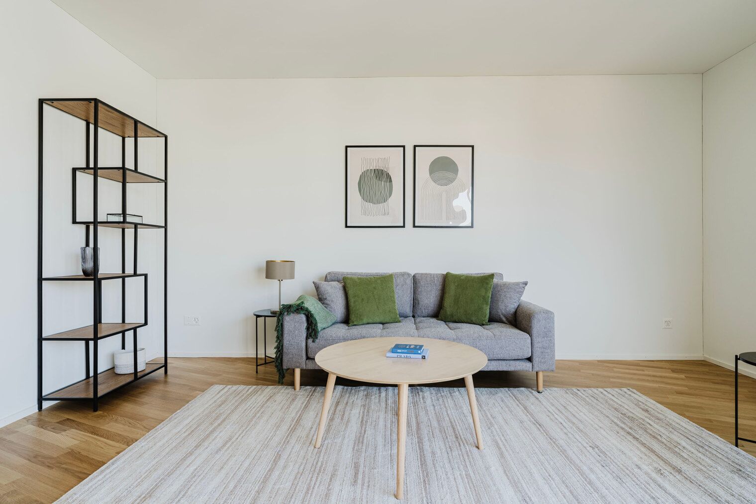Un salon avec un canapé gris, une table basse ronde, une étagère en métal noir et deux œuvres d’art abstraites encadrées accrochées au mur.