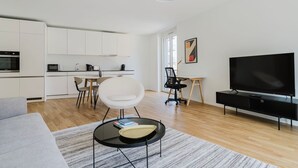 A modern living room with a flat-screen TV, a dining area with a white table and chairs, a kitchen with white cabinets, and a grey sofa.