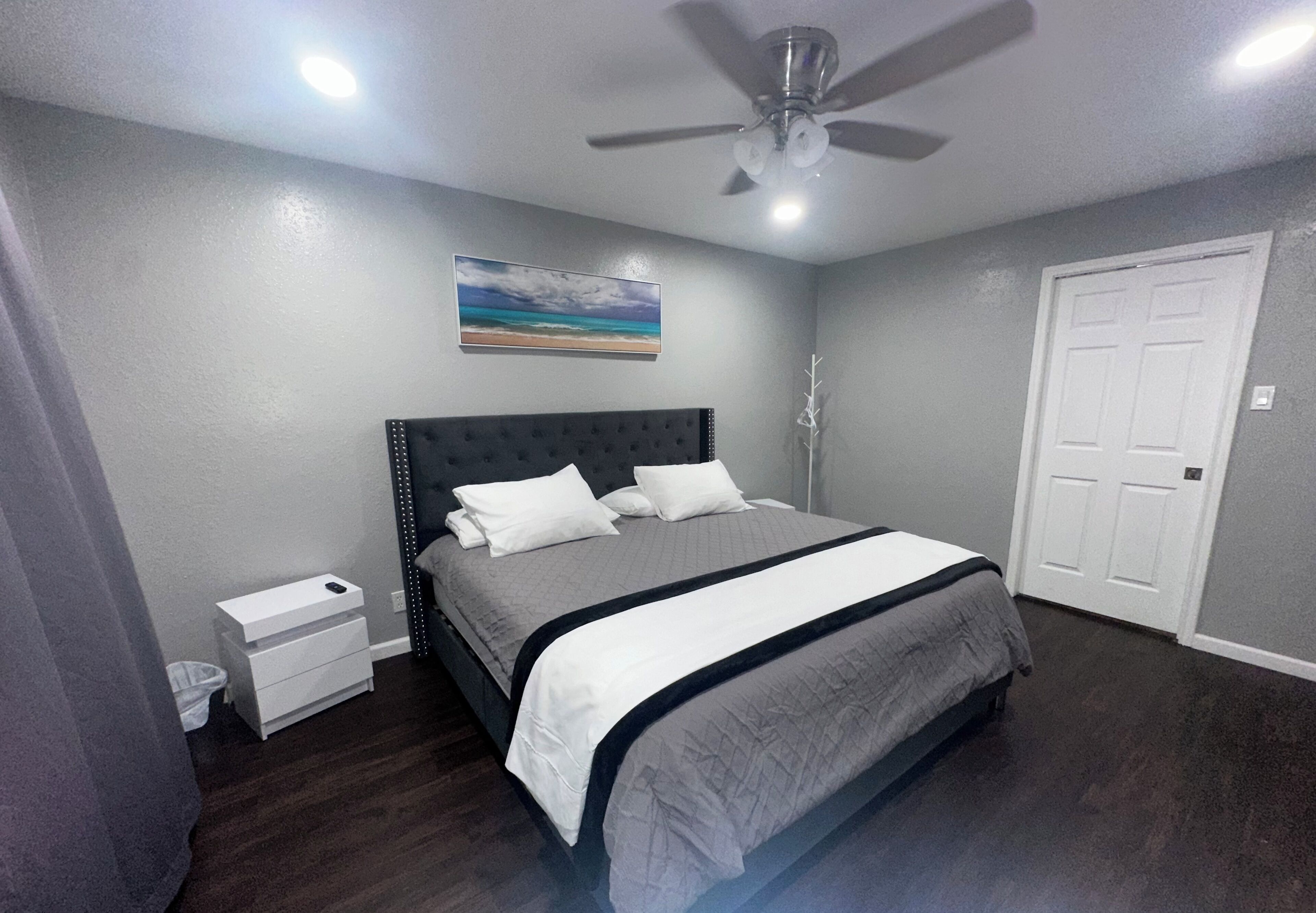 A bedroom with a bed, a nightstand, a ceiling fan, a door, and a painting on the wall.
