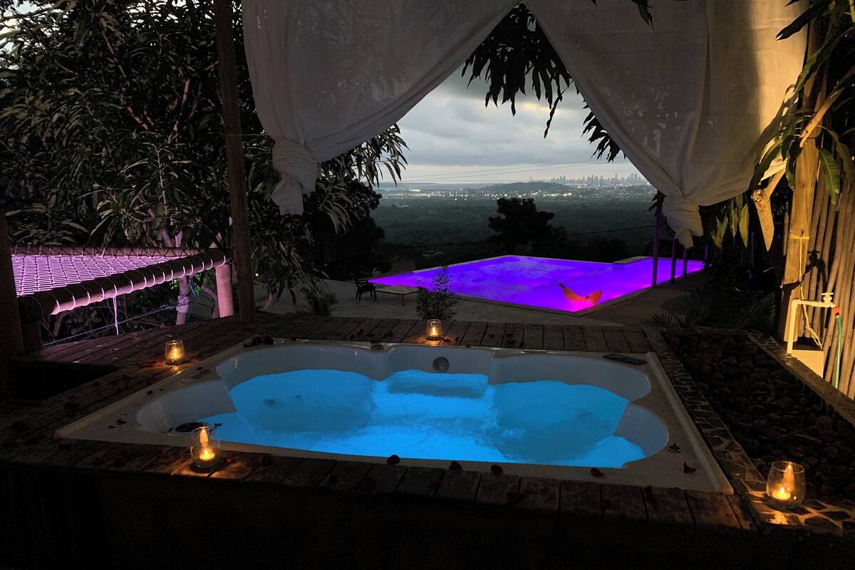 Uma banheira de hidromassagem com vista para o horizonte urbano, rodeada por vegetação e uma pérgola com iluminação roxa.