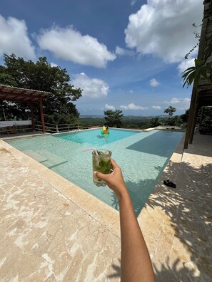 A swimming pool with clear water, a person holding a drink, and a view of the city in the background. - Ada Marina (Turbaco)
