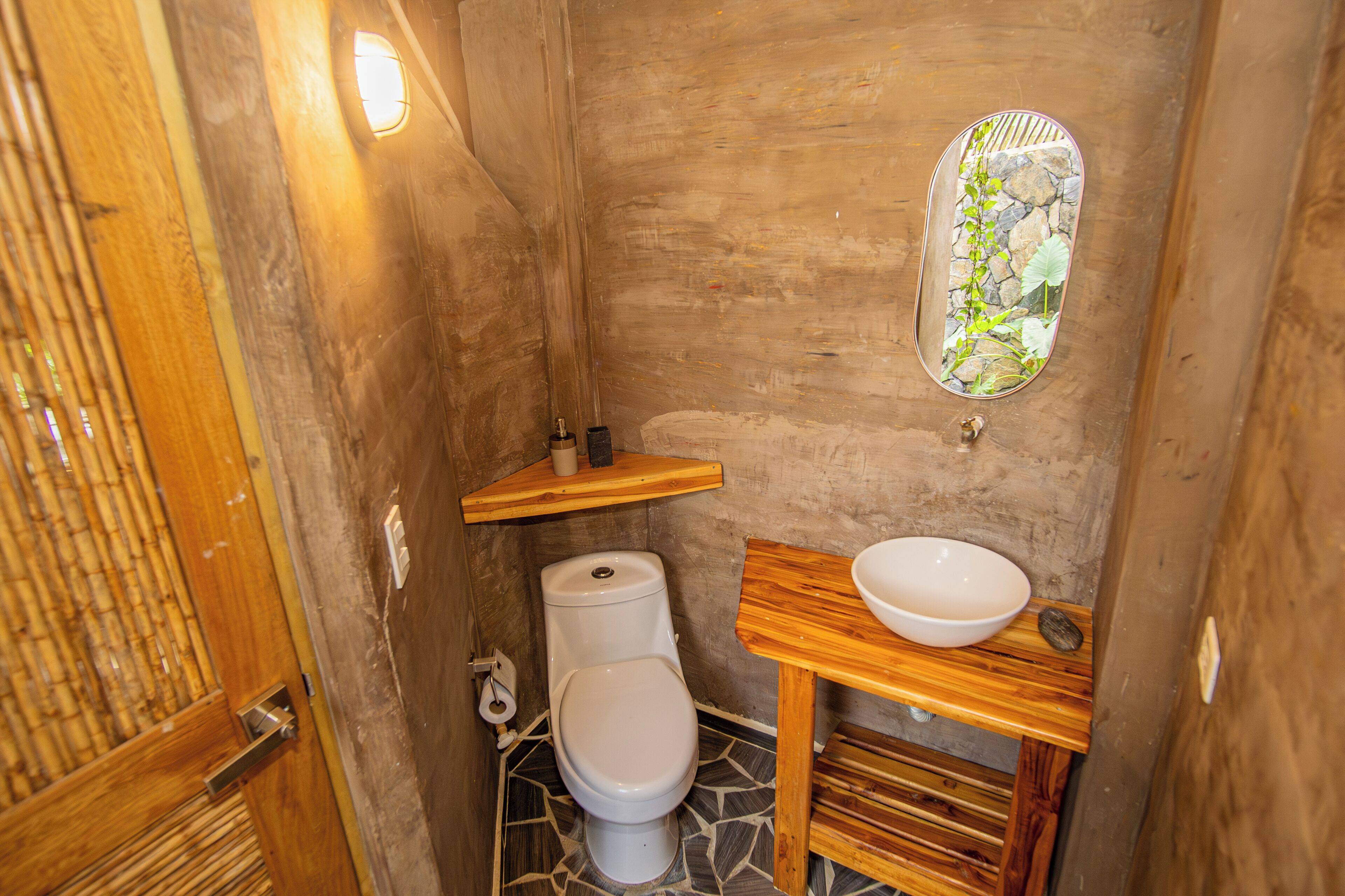 A compact bathroom with a toilet, a wooden vanity with a bowl sink, a mirror, and a bamboo door.