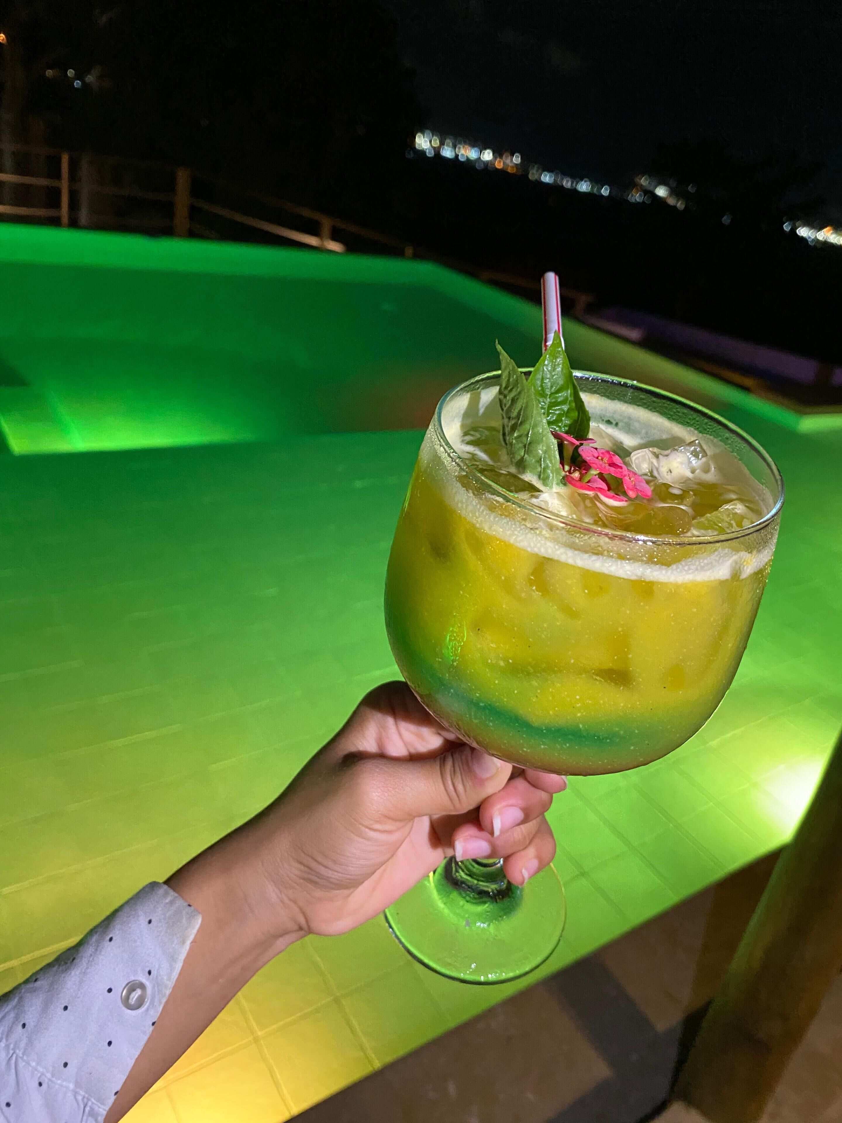 A hand holding a cocktail with a lime garnish, set against a green-lit background.