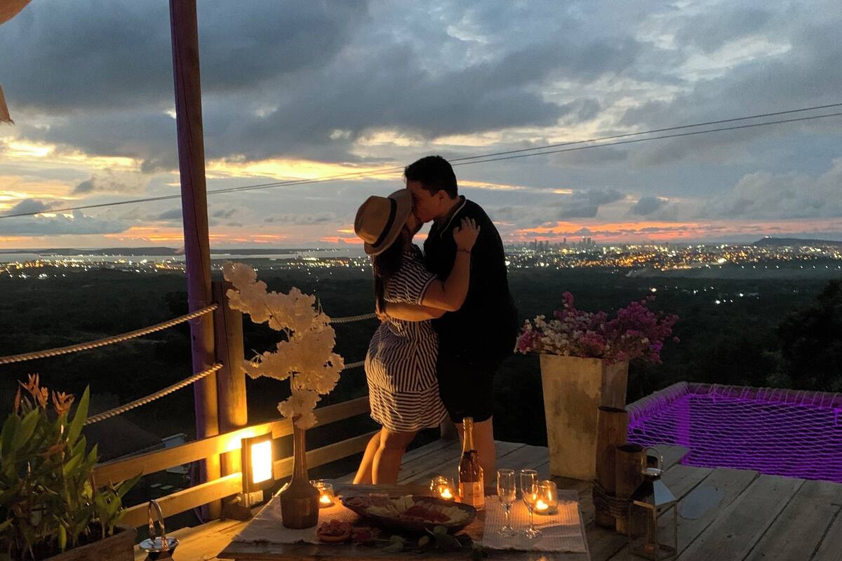 Um casal se abraçando em um deck de madeira com vista para a cidade ao pôr do sol.