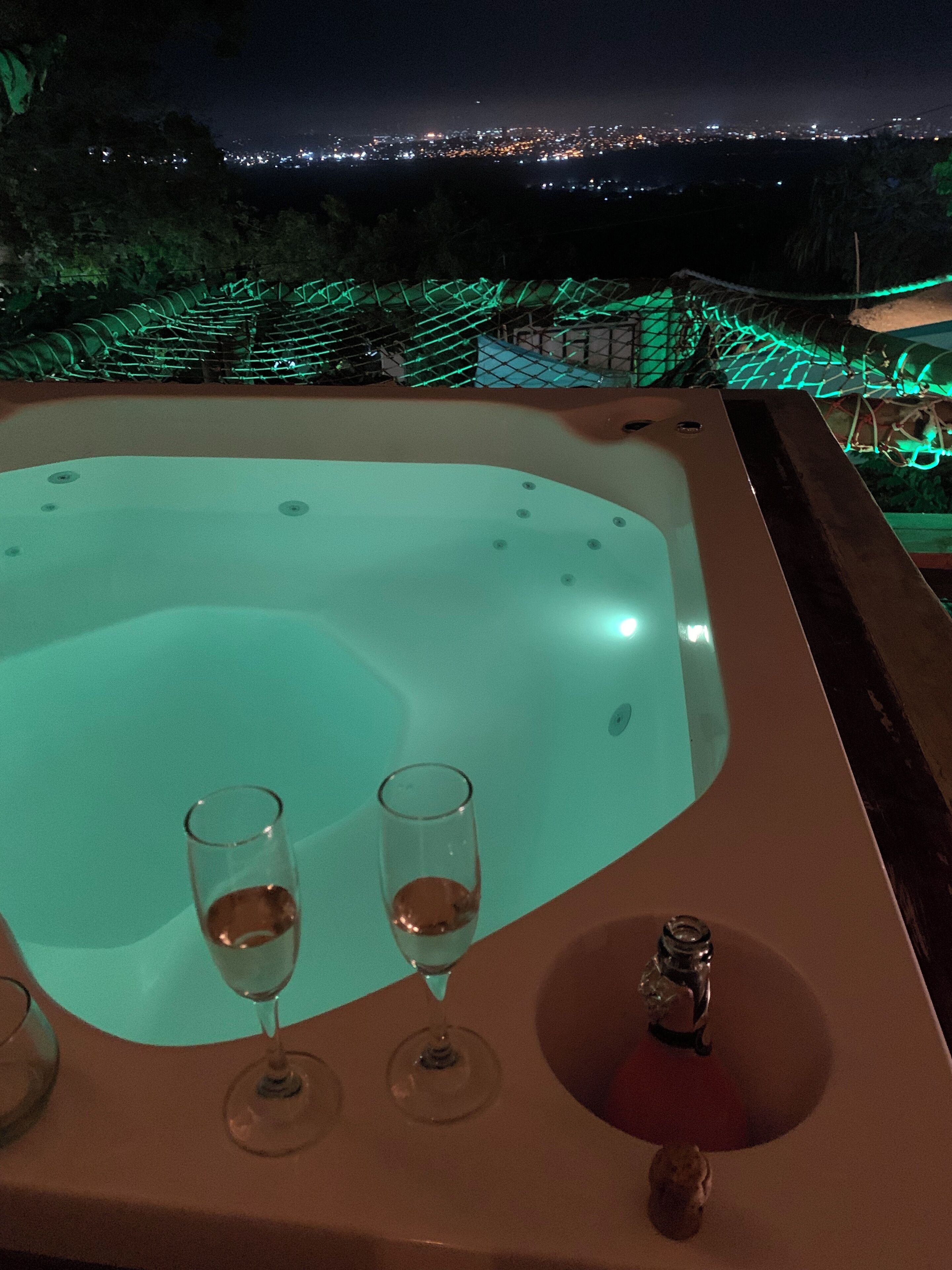 Un jacuzzi con dos copas de vino sobre una mesa, con vistas a un paisaje urbano nocturno.