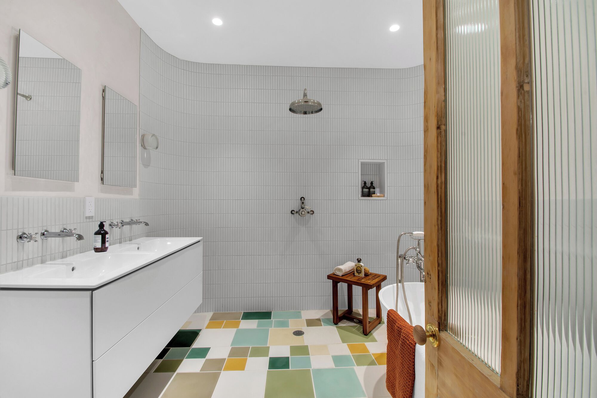 A modern bathroom with a white tiled shower, a sink with a mirror, and a small wooden stool.