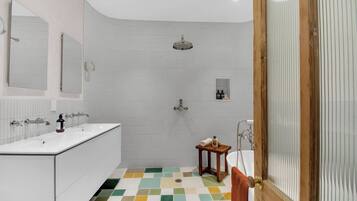 A modern bathroom with a white tiled shower, a sink with a mirror, and a small wooden stool.