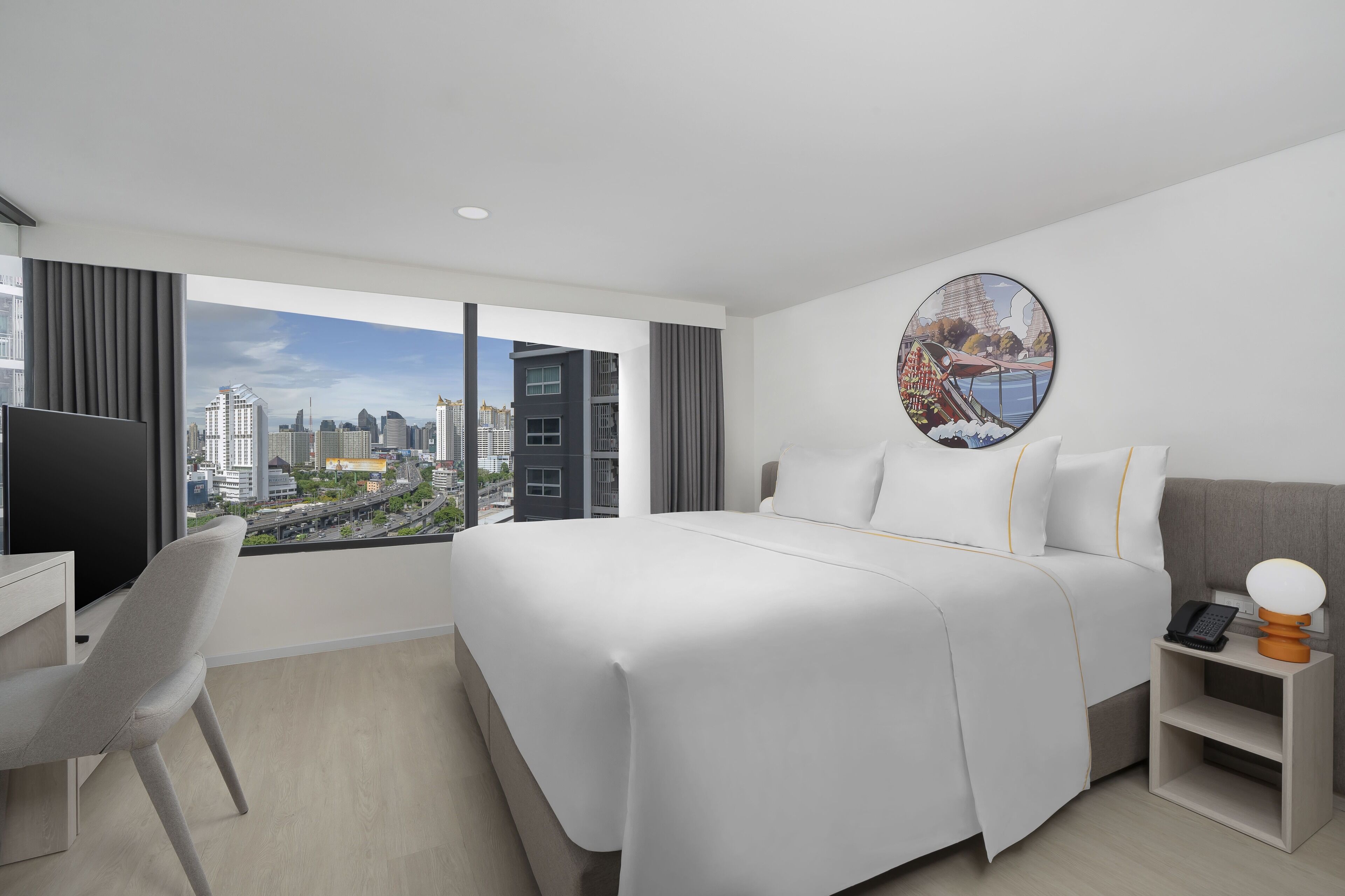 Une chambre d’hôtel moderne avec un grand lit, un bureau et une chaise. La fenêtre offre une vue sur la ville, et un tableau rond est accroché au mur.