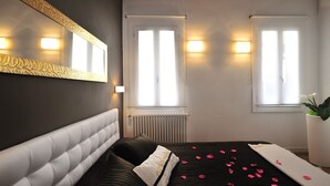 A bedroom with a large mirror, two windows with curtains, a radiator, and a bed with rose petals. - Dimora San Zanipolo by Wonderful Italy (Venice)