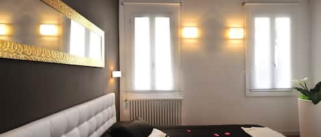 Une chambre avec un grand miroir, deux fenêtres avec des rideaux, un radiateur et un lit parsemé de pétales de rose.