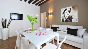 A modern living room with a white sofa, a glass dining table with pink flower petals, white chairs, a flat-screen TV, and a painting on the wall. - Dimora San Zanipolo by Wonderful Italy (Venice)