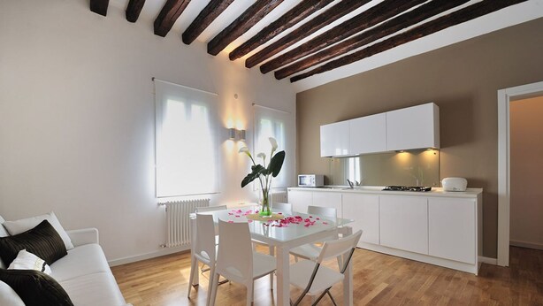 A modern living room with a glass dining table, white furniture, and a kitchen area with white cabinets and a stove. - Dimora San Zanipolo by Wonderful Italy (Venice)