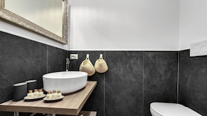 Een moderne badkamer met een grote witte wastafel, donkere betegelde wanden, een houten wastafelmeubel en een wit toilet.