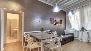 A modern living room with a dining table, white chairs, a grey sofa, and a large window with sheer curtains. - San Marco Promenade by Wonderful Italy (Venice)
