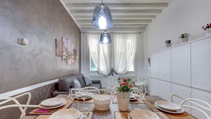 A dining area with a table set for six, white chairs, a hanging light fixture, and a view of a cityscape through a window with sheer curtains. - San Marco Promenade by Wonderful Italy (Venice)
