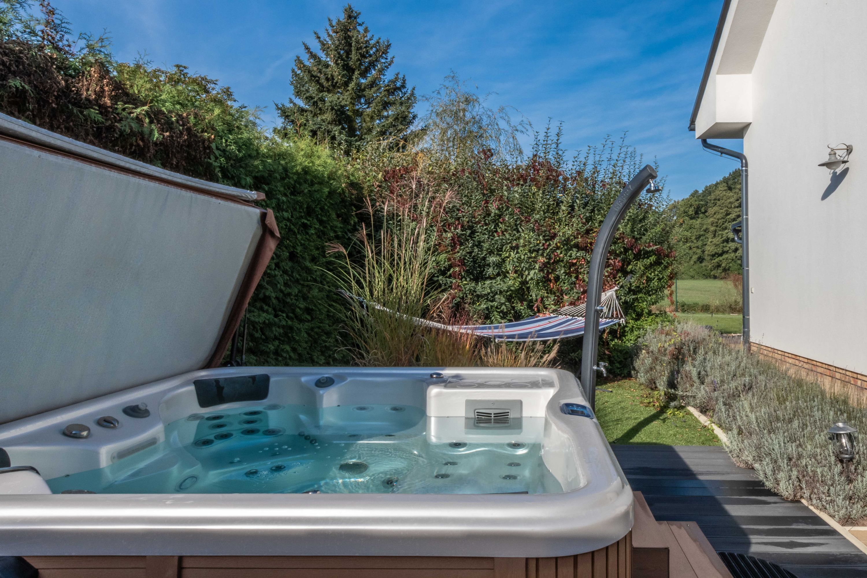 A hot tub with a wooden deck, surrounded by greenery and a white building.