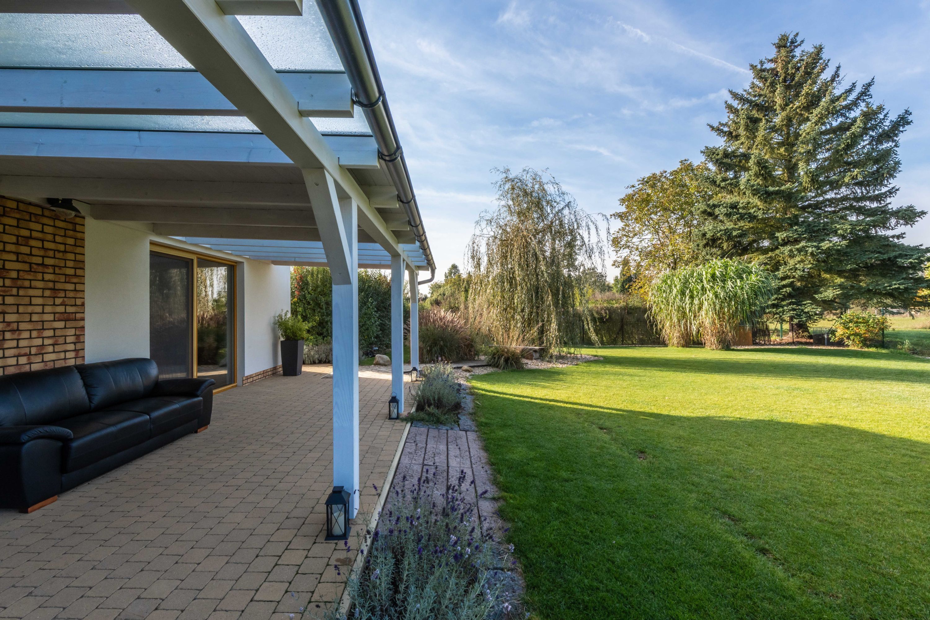 A covered patio area with a black leather sofa, a brick wall, and a glass door leading to a garden with a lawn and trees.