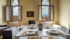 Un comedor con una mesa dispuesta para cuatro, dos ventanas con contraventanas abiertas, un cuadro enmarcado y un radiador.
