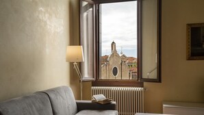 Una habitación con un sofá gris, una lámpara de lectura, un radiador y una ventana con vista a un edificio.