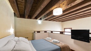 Un dormitorio con cama, un televisor de pantalla plana montado en la pared, vigas de madera en el techo y una ventana.