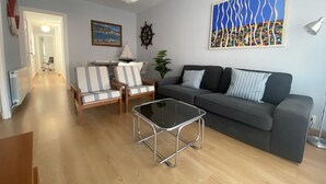 Ένα σαλόνι με ένα ν καναπέ, δύο καρέκλες με ριγέ ύφασμα, ένα τραπεζάκι σαλονιού από γυαλί, ένα μοντέλο ιστιοπλοϊκού σκάφους, ένα πηδάλιο πλοίου και έναν πολύχρωμο αφηρημένο πίνακα.