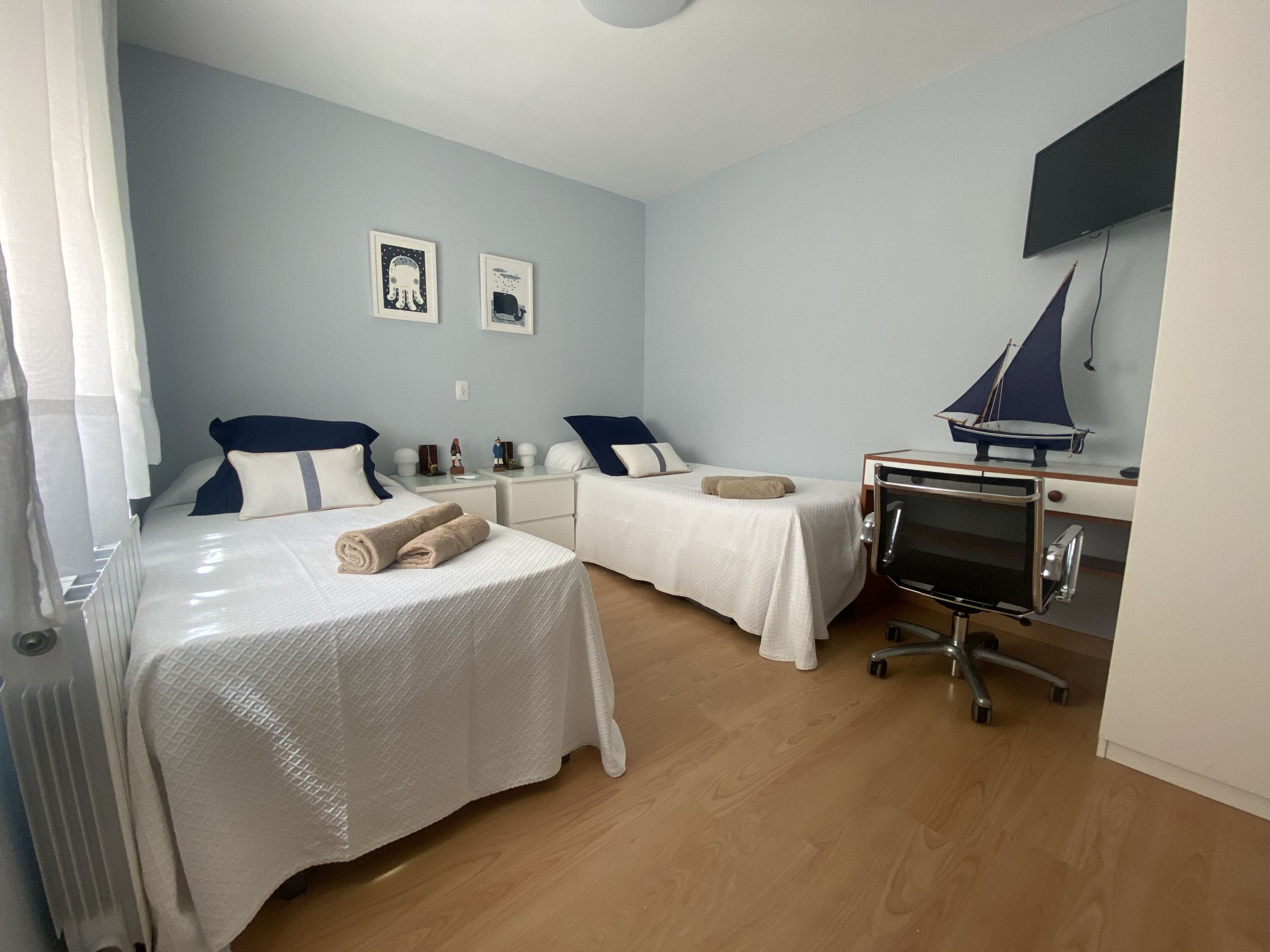 A bedroom with two beds, a desk with a computer, and a sailboat model on the wall.