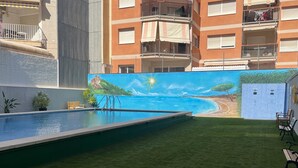 Μια πισίνα με μια τοιχογραφία που απεικονίζει μια παραλία, μια καταπράσινη περιοχή και ένα κτίριο με μπαλκόνια.