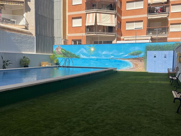 A swimming pool with a mural of a beach scene, a grassy area, and a building with balconies.