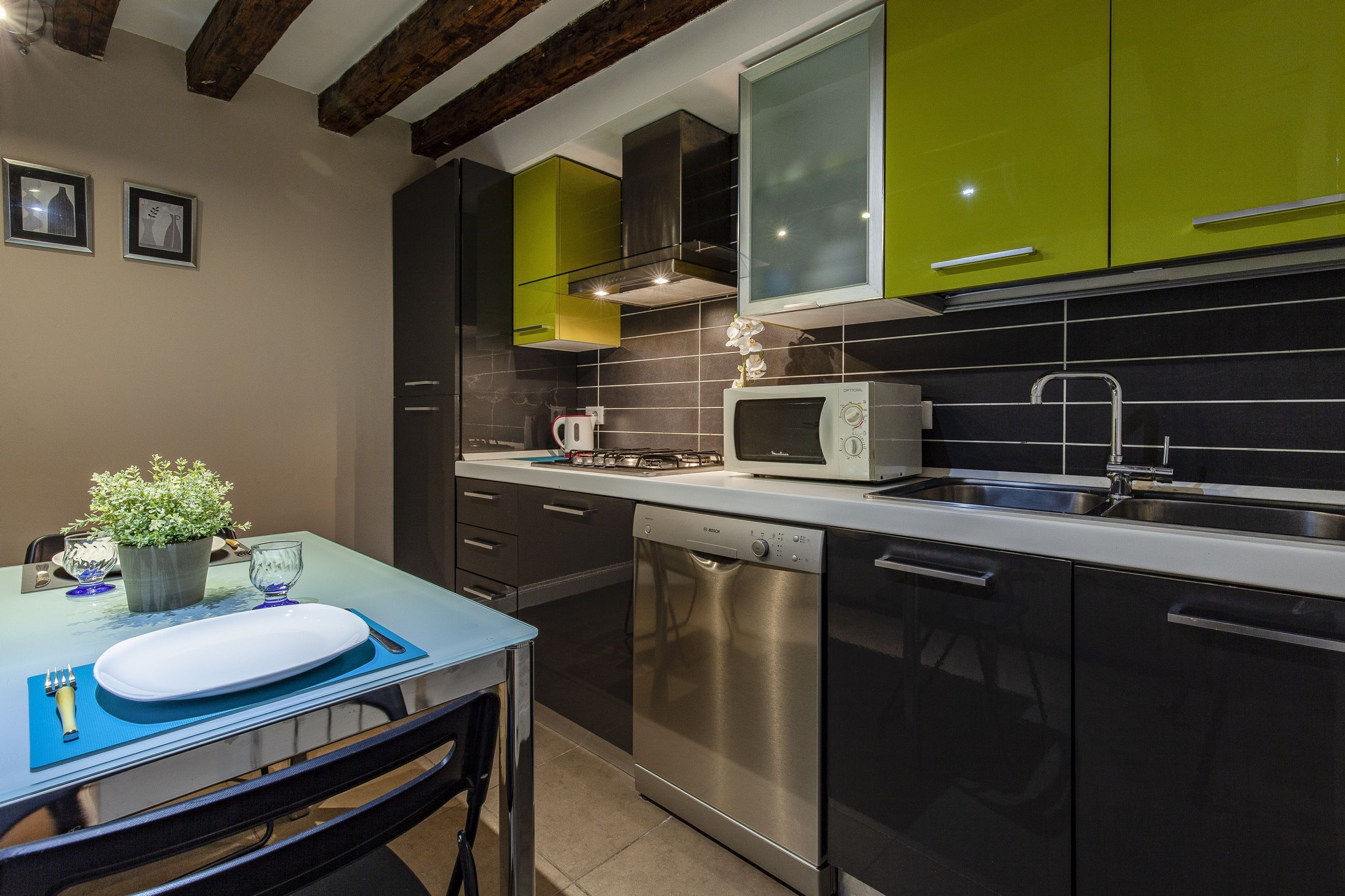 A modern kitchen with stainless steel appliances, a dining table with a blue placemat, and green cabinets.