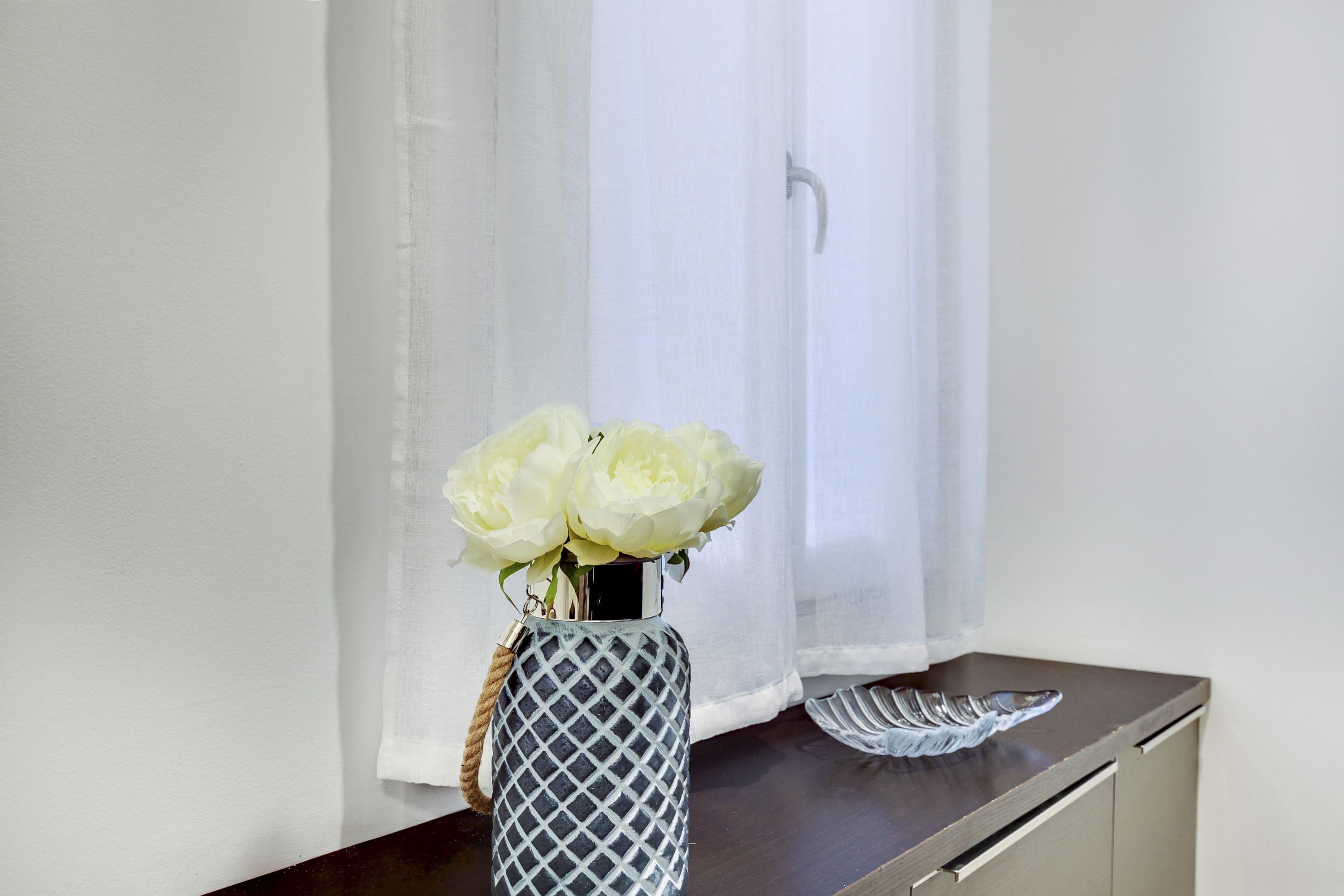 A vase with white flowers on a wooden surface near a window with sheer curtains.
