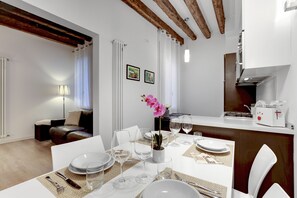Uma sala de jantar com uma mesa branca posta para quatro pessoas, incluindo pratos, copos e talheres. Há uma orquídea em vaso sobre a mesa, e a cozinha apresenta um design moderno, com forno embutido e micro-ondas.