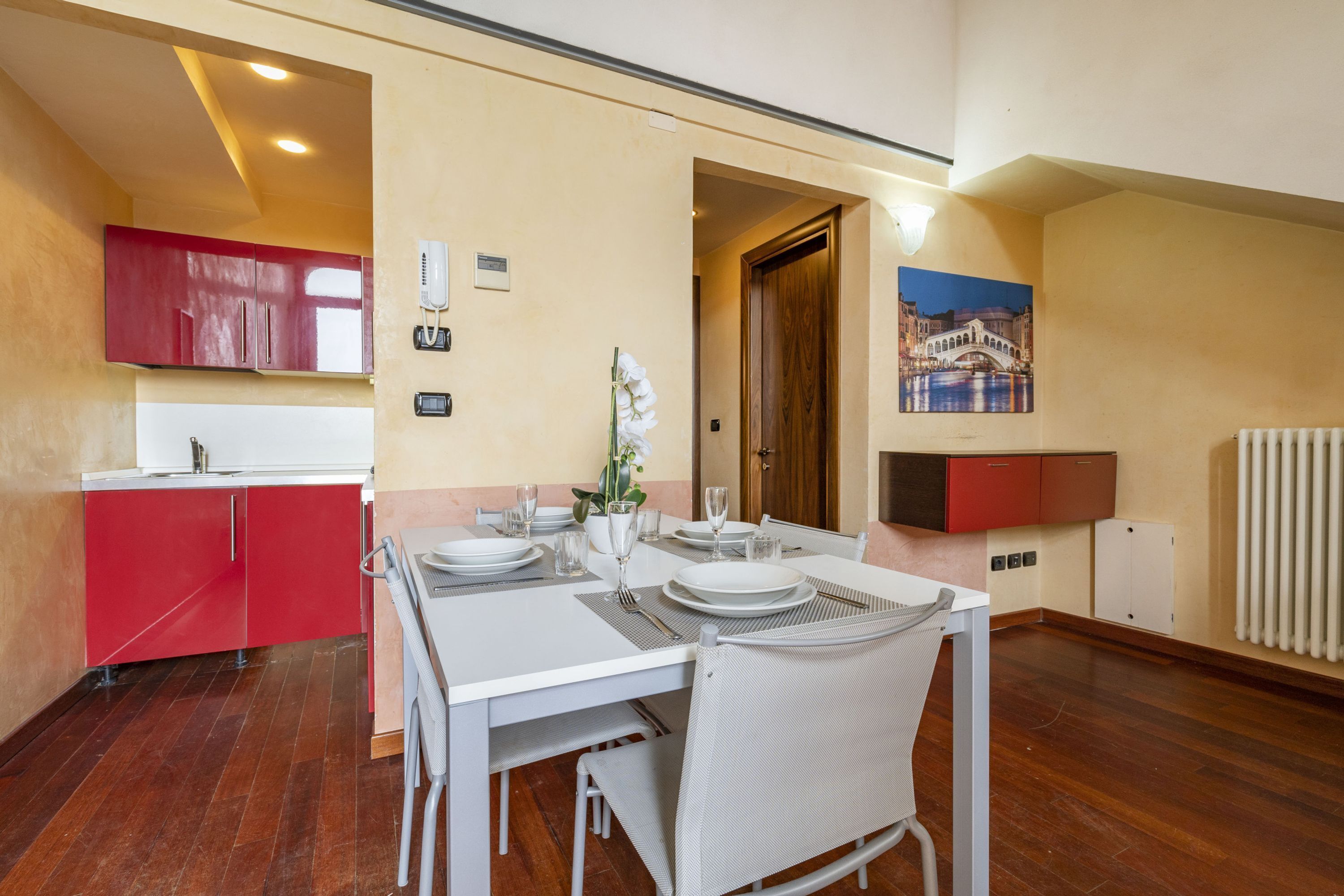 A dining area with a white table set for two, red cabinets, a painting of a bridge, and a radiator.