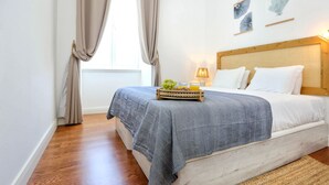 A bedroom with a bed, a bedside table with a tray of fruit and juice, a lamp, and a window with curtains.