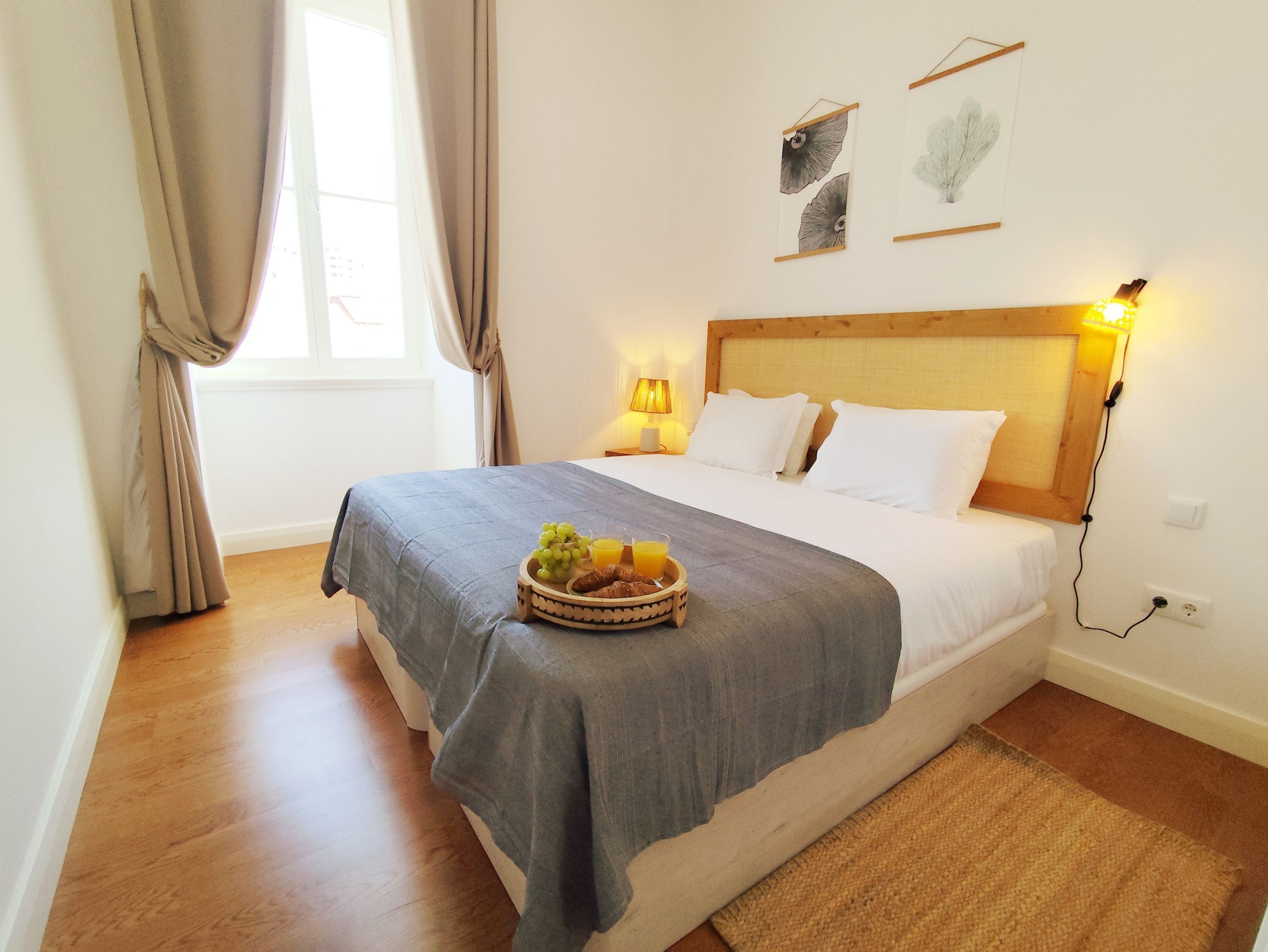 Una cama bien hecha con un edredón gris, una mesita de noche con una lámpara y una cesta de frutas sobre la cama.