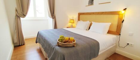 Una cama bien hecha con un edredón gris, una mesita de noche con una lámpara y una cesta de frutas sobre la cama.
