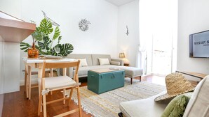A modern living room with a wooden dining table, a sofa, a teal ottoman, and a large plant.