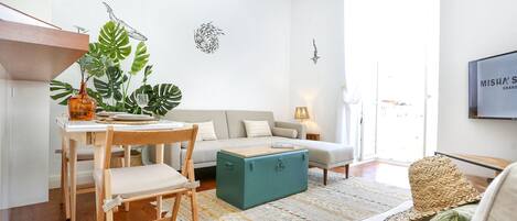 A modern living room with a wooden dining table, a sofa, a teal ottoman, and a large plant.