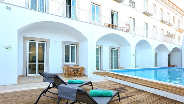 An outdoor swimming pool area with lounge chairs and a table, surrounded by white building with arched windows and balconies.